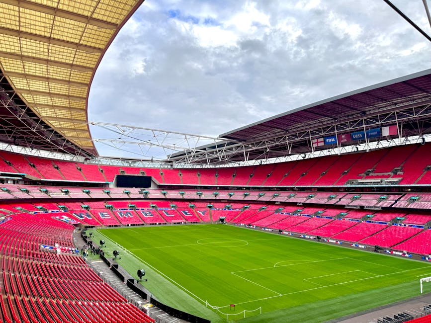An aerial view of a large, modern football stadium with a bright green pitch, surrounded by a white curved roof structure and steel support beams. The stadium is situated in a suburban area with well-
